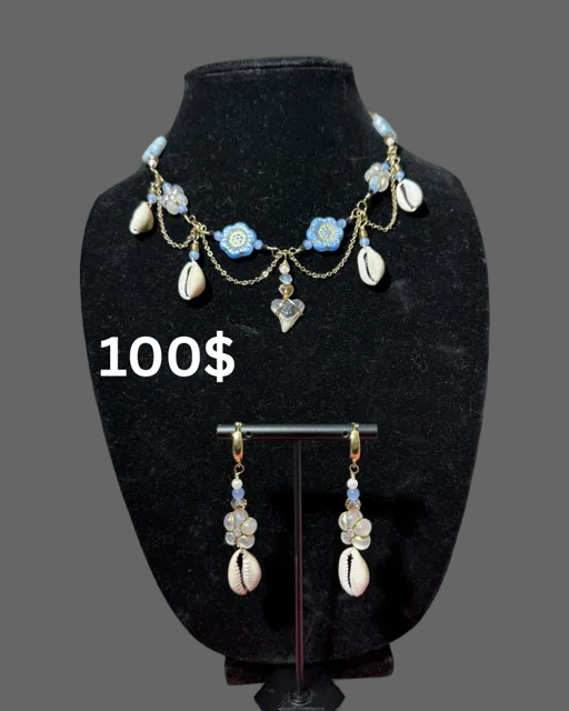 Boho-style jewelry set featuring a necklace and matching drop earrings displayed on a black velvet mannequin bust. The jewelry has light blue floral charms, white cowrie shells, and gold-toned chains and hooks.